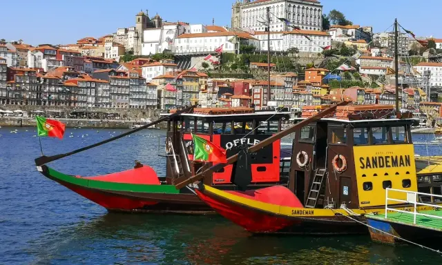 Tradycyjne łodzie Rabelo cumują w malowniczym porcie miasta Porto, z kolorowymi domami na wzgórzu w tle.