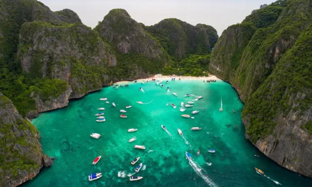 Maya Bay tętni życiem, pełna łodzi i turystów na piaszczystej plaży, otoczona stromymi, porośniętymi zielenią klifami.