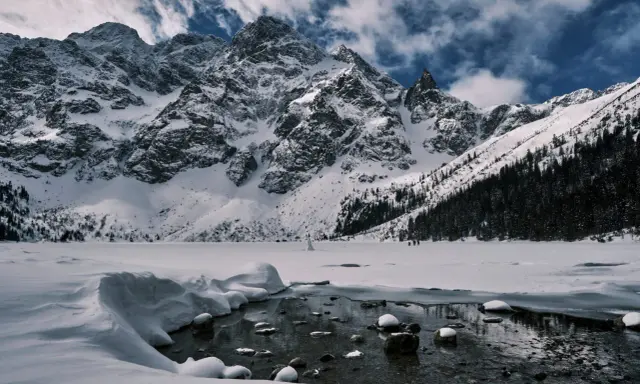 Morskie Oko zimową porą. Lodowate wody jeziora odbijają ośnieżone szczyty Tatr, a na horyzoncie widać ludzi spacerujących po zamarzniętej tafli.