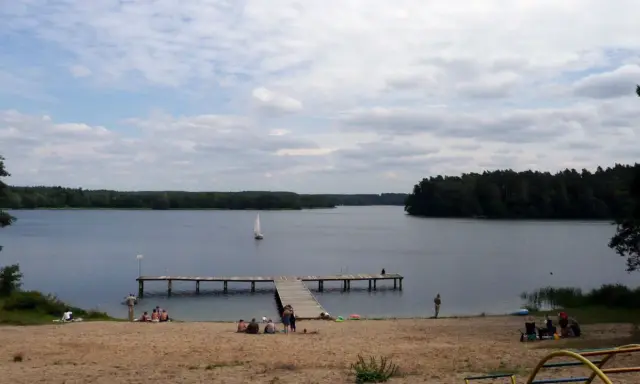 Spokojny dzień na plaży nad jeziorem, żaglówka na wodzie, drewniane molo. Pojezierze Brodnickie zaprasza do wypoczynku.