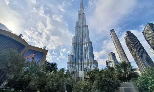 Burj Khalifa, najwyższy budynek w Dubaju, wznosi się dumnie na tle błękitnego nieba z chmurami. Otoczony zielenią i innymi drapaczami chmur.
