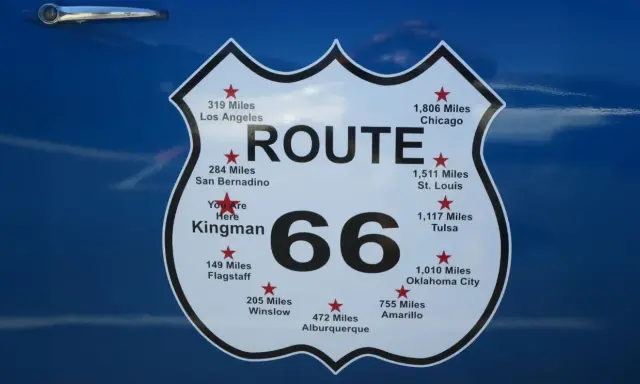 Tablica z napisem "ROUTE 66" i odległościami do miast, np. Chicago, Los Angeles, St. Louis.