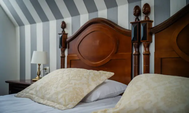 Eleganckie łóżko z drewnianym zagłówkiem i ozdobnymi poduszkami. Ciekawe, ile wynosi ryczałt za nocleg w takim miejscu?