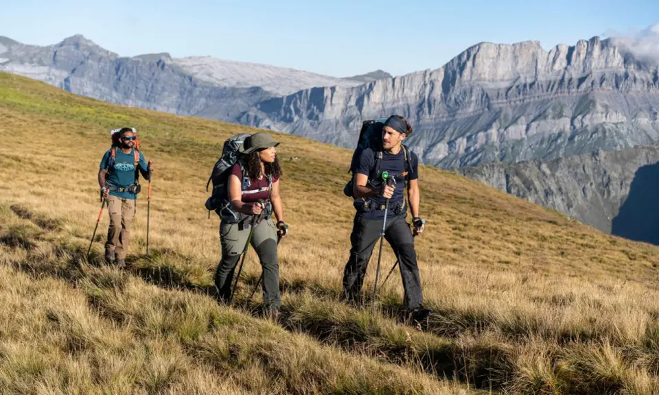 Trzy osoby z plecakami i kijkami trekkingowymi idące po trawiastym zboczu górskim.