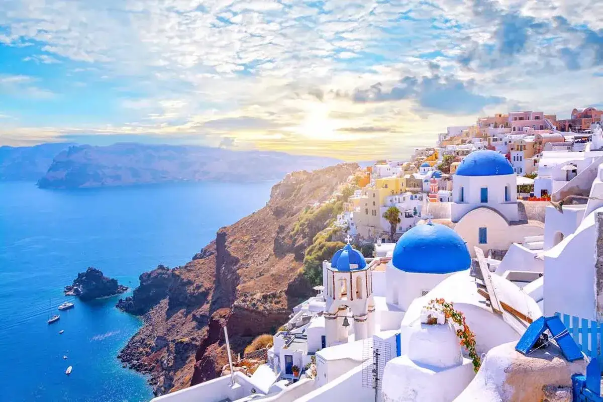 Santorini wakacje: białe domy z niebieskimi kopułami na klifie nad lazurowym morzem, w tle góry i słońce.