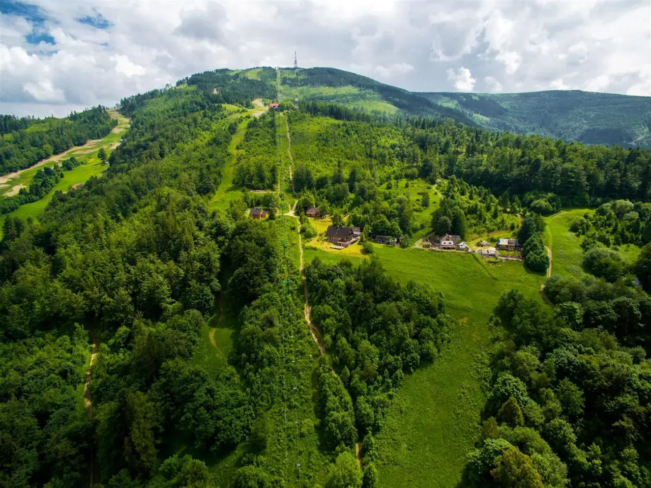 Widok na najwyższy szczyt Beskidu Śląskiego, Skrzyczne, z kolejką linową i kilkoma domkami pośród zielonych zboczy.