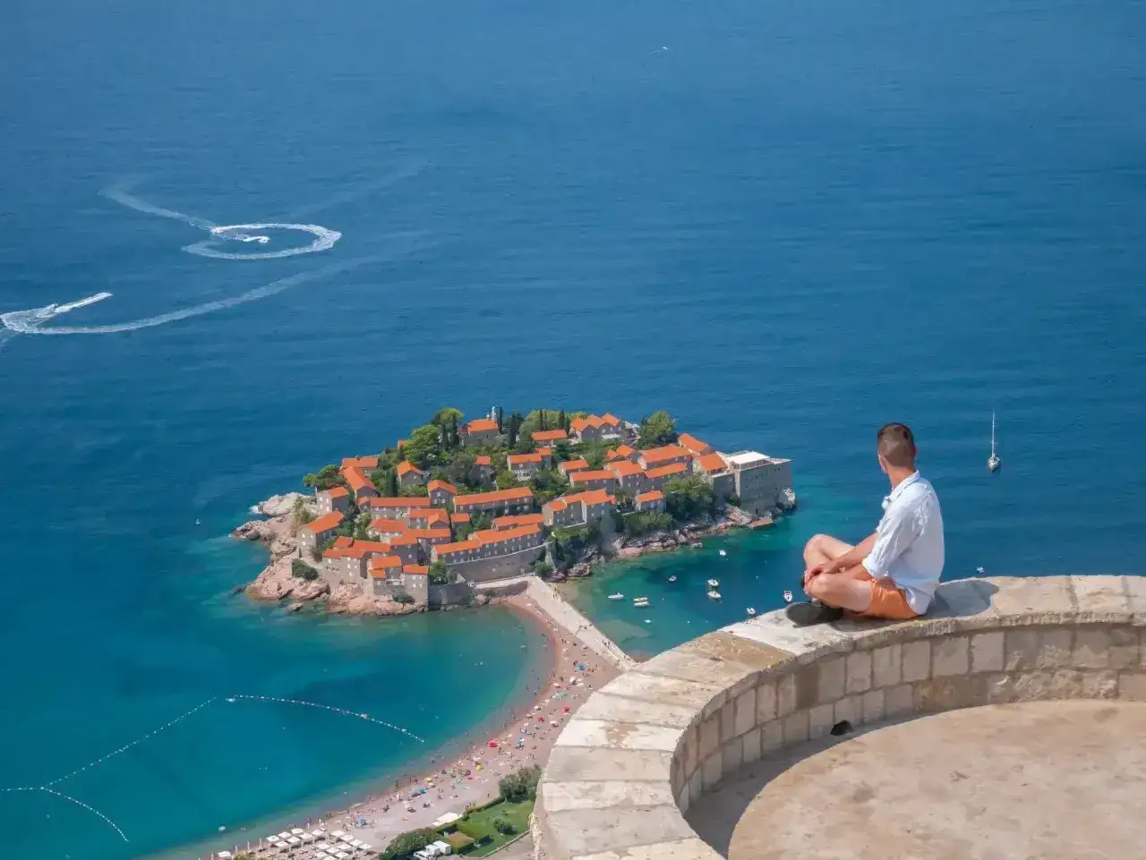 Czarnogóra na co uważać: widok na wyspę Sveti Stefan, turysta siedzi na murku, podziwiając panoramę.