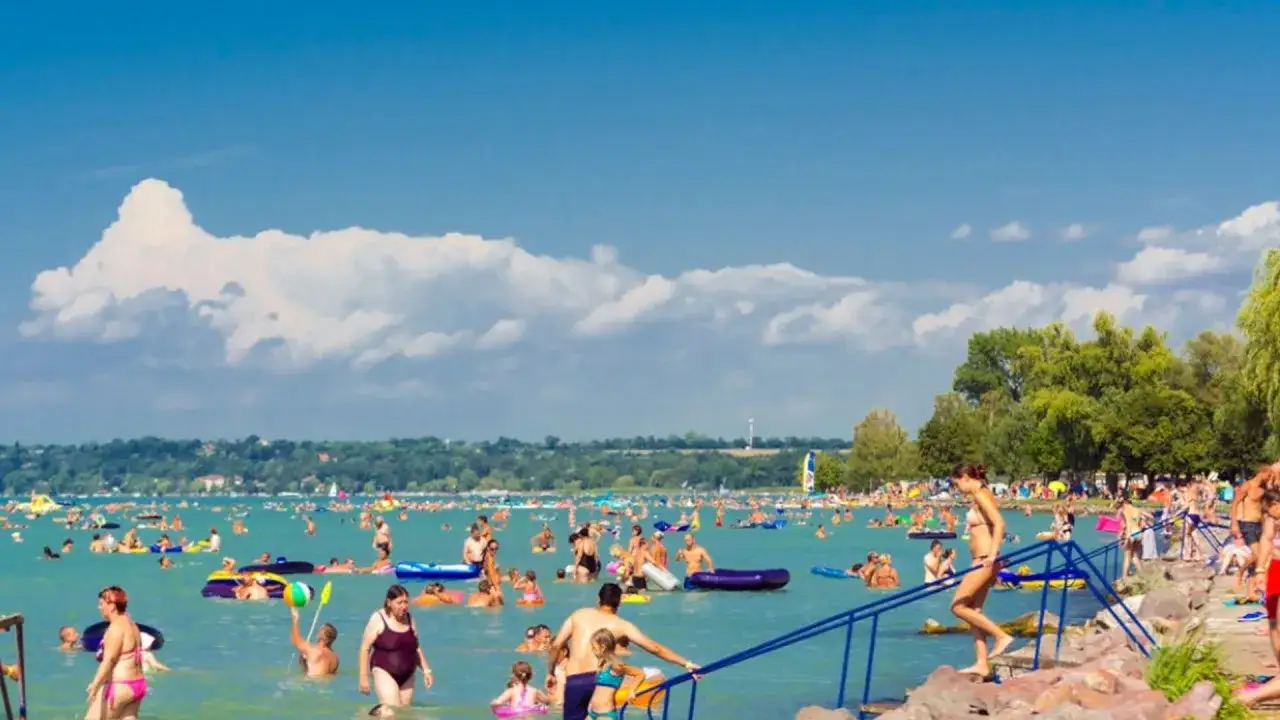 Tłum ludzi cieszy się słońcem i wodą na zatłoczonej balaton plaży. Wiele osób pływa, relaksuje się na materacach i bawi się w wodzie.