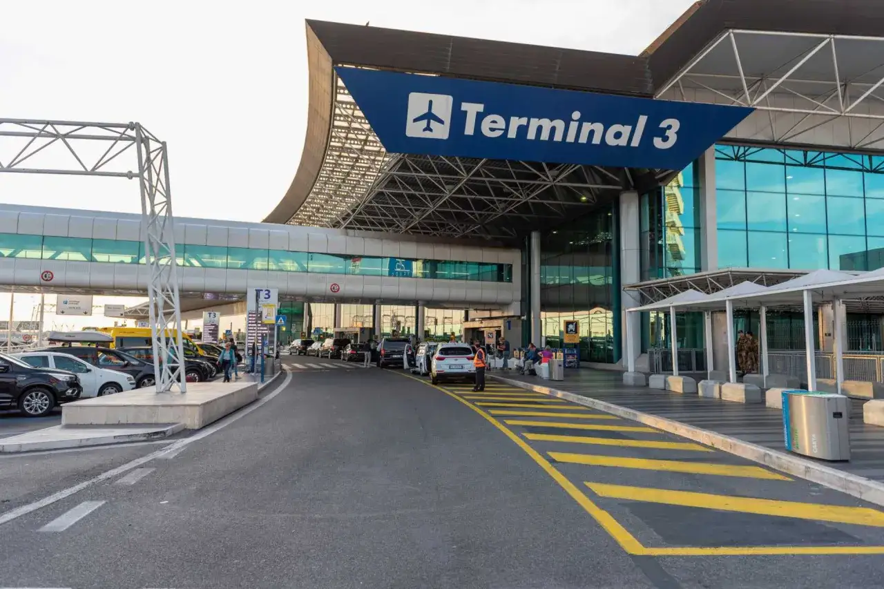 Wejście do Terminalu 3 lotniska Fiumicino. Samochody czekają przed budynkiem, a pasażerowie z bagażami kierują się do środka.
