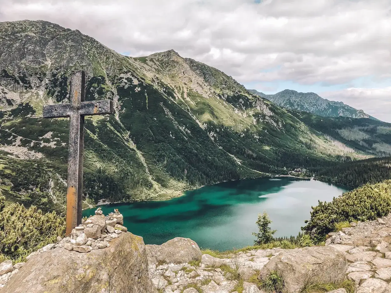 Kamienny krzyż na pierwszym planie, za nim turkusowe Morskie Oko i zielone zbocza Tatr od strony polskiej.