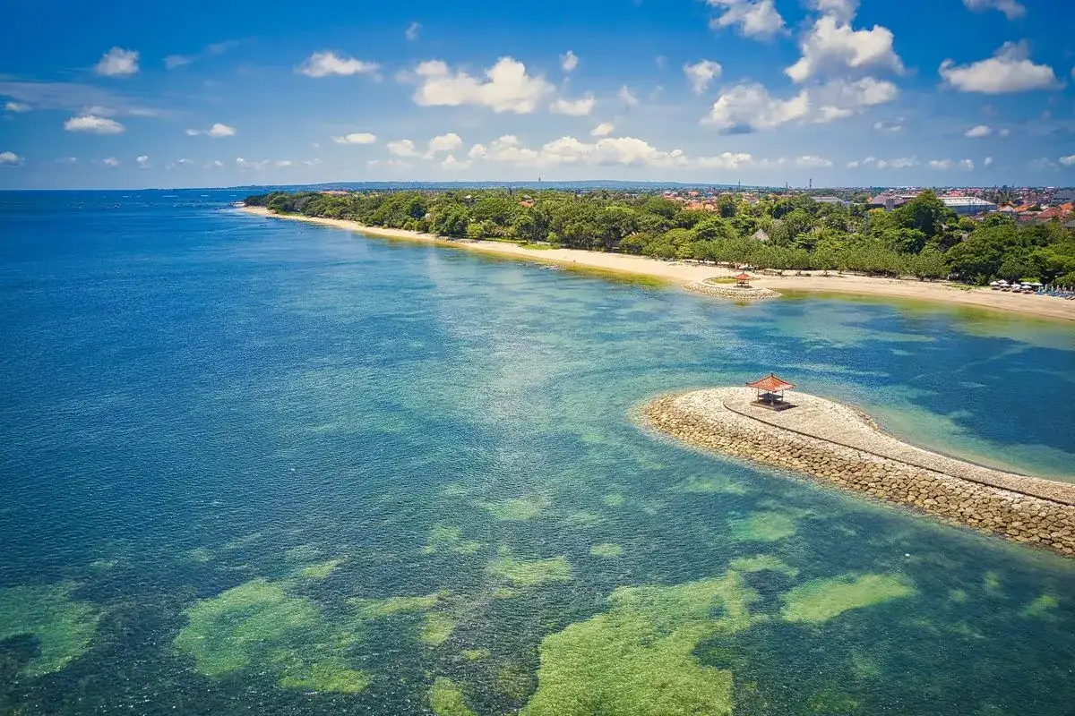 Spokojne wody Sanur, z kamiennym molem prowadzącym do pawilonu, plażą i bujną zielenią w tle.
